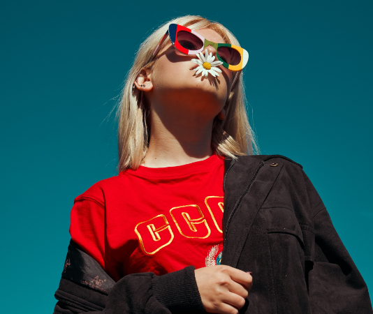 Blonde Person mit farbenfrohen Sonnenbrillen und Blüte im Mund vor klarem, blauem Himmel.